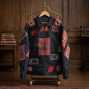 Puede incluir: Una chaqueta negra con cremallera que presenta un patrón de parches dispersos con motivos rectangulares y cuadrados en tonos rojo, azul y dorado. La prenda tiene un cuello clásico, mangas largas y un cierre de cremallera central en una percha.