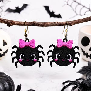 May include: A pair of dangling earrings featuring black spider charms with large white eyes, pink cheeks, and a bright pink bow on top. The spiders have eight legs and are attached to gold-toned metal earring hooks for pierced ears.