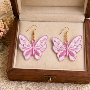 May include: A pair of butterfly earrings with pink and white embroidered wings, displayed in a wooden jewelry box. The earrings have gold-colored hooks and a delicate design, perfect for adding a touch of whimsy to any outfit.