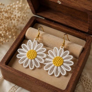 Peut inclure: Une paire de boucles d'oreilles pendantes représentant des motifs de fleurs de marguerite brodées en blanc avec des cœurs jaunes. Les pétales présentent un délicat motif en maille ajourée et sont fixés à des crochets en métal doré.