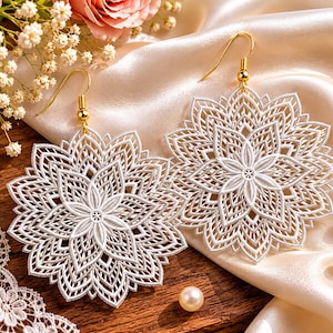 Könnte beinhalten: Ein Paar weiße Spitzenohrringe mit einem detaillierten, symmetrischen, mandalaartigen Blumenmuster. Jeder Ohrring hängt an einem goldfarbenen Metallhaken. Das filigrane, durchbrochene Design bildet eine zarte, sternförmige Struktur mit mehreren Schichten von Blütenblättern, die von der Mitte ausstrahlen.