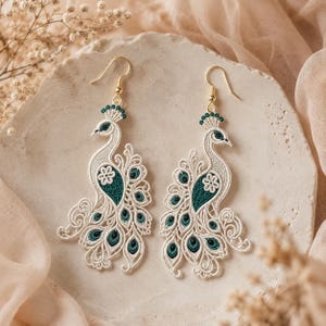 May include: A pair of drop earrings featuring intricate white lace embroidery in the shape of peacocks. The peacock bodies are accented with deep teal thread details and small floral motifs, suspended from gold-toned metal earring hooks.