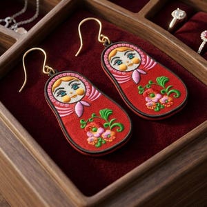 May include: A pair of red embroidered Matryoshka doll earrings with gold-colored hooks. The earrings feature a doll face with blue eyes and a pink scarf, and are decorated with floral embroidery. The earrings are displayed in a wooden jewelry box.