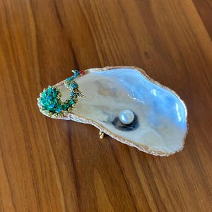 May include: A decorative oyster shell dish with a gold-painted rim, featuring a peacock-shaped embellishment adorned with green and blue crystals on one side and a single white spherical pearl resting in the center of the iridescent interior.