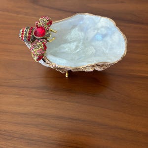 May include: A decorative, iridescent white shell-shaped dish with a gold-colored rim. A jeweled bee with red and gold accents adorns the side. The dish is set on a wooden surface.