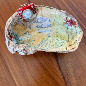 May include: A decorative oyster shell dish with a gold-painted rim. The interior features vintage paper with blue script and floral accents. A red camera-shaped charm with a pearl and chain is attached to the dish.
