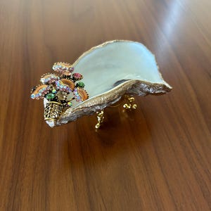 May include: A decorative oyster shell dish featuring a gold-painted rim and base. The interior is smooth and white. Attached to one side is a cluster of metallic, multi-colored rhinestone-encrusted mushroom figures in shades of pink and green.