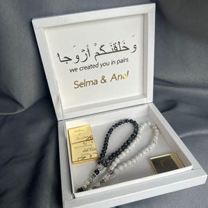 Könnte beinhalten: Eine weiße Schatulle mit goldener Aufschrift „we created you in pairs“ und den Namen „Selma & Anel“. Enthält zwei Perlenketten, eine schwarze und eine weiße, sowie zwei goldfarbene rechteckige Gegenstände.