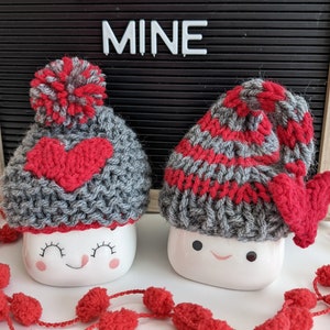 Puede incluir: Dos tazas de cerámica blanca con caras sonrientes, que llevan gorros de punto grises y rojos con pompones. Las tazas están sobre una superficie blanca con una guirnalda de pompones rojos. Un tablero de letras negro detrás de las tazas dice "BE MINE".