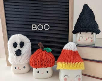 Puede incluir: Cuatro tazas de cerámica blanca con caras sonrientes, cada una adornada con un sombrero de punto con temática de Halloween. Los sombreros incluyen un fantasma, una calabaza, un caramelo de maíz y un sombrero de bruja. Una pizarra negra muestra la palabra "BOO".