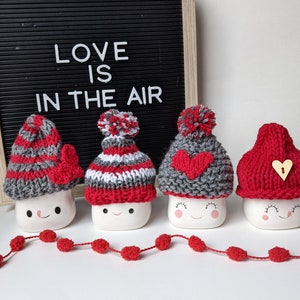 Puede incluir: Cuatro tazas de cerámica blanca con gorros de punto rojos y grises. Las tazas tienen ojos y sonrisas pintadas. Los gorros tienen pompones y corazones. Una guirnalda de pompones rojos está delante de las tazas. Un tablero de letras negro detrás de las tazas dice "LOVE IS IN THE AIR".