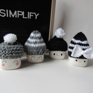 Puede incluir: Cuatro figuras de malvavisco de cerámica con gorros de punto con pompones y lazos. Los gorros son de varios colores: gris, negro y blanco. Los malvaviscos tienen caras sonrientes con mejillas rosadas. La palabra "SIMPLIFY" se muestra sobre un fondo negro.