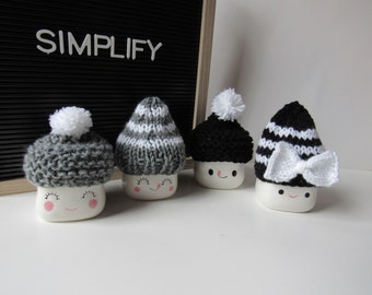 Könnte beinhalten: Vier Keramik-Marshmallow-Figuren mit gestrickten Mützen mit Bommeln und Schleifen. Die Mützen sind in verschiedenen Farben: Grau, Schwarz und Weiß. Die Marshmallows haben lächelnde Gesichter mit rosa Wangen. Das Wort "SIMPLIFY" wird auf schwarzem Hintergrund angezeigt.