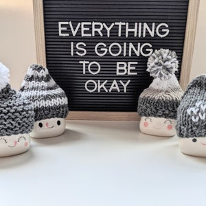 K&ouml;nnte beinhalten: Vier wei&szlig;e Keramikbecher mit fr&ouml;hlichen Gesichtern tragen gestrickte M&uuml;tzen. Die M&uuml;tzen sind grau und wei&szlig; mit Bommeln. Die Becher stehen auf einer wei&szlig;en Oberfl&auml;che vor einem schwarzen Buchstabenbrett mit der Aufschrift "Alles wird gut."