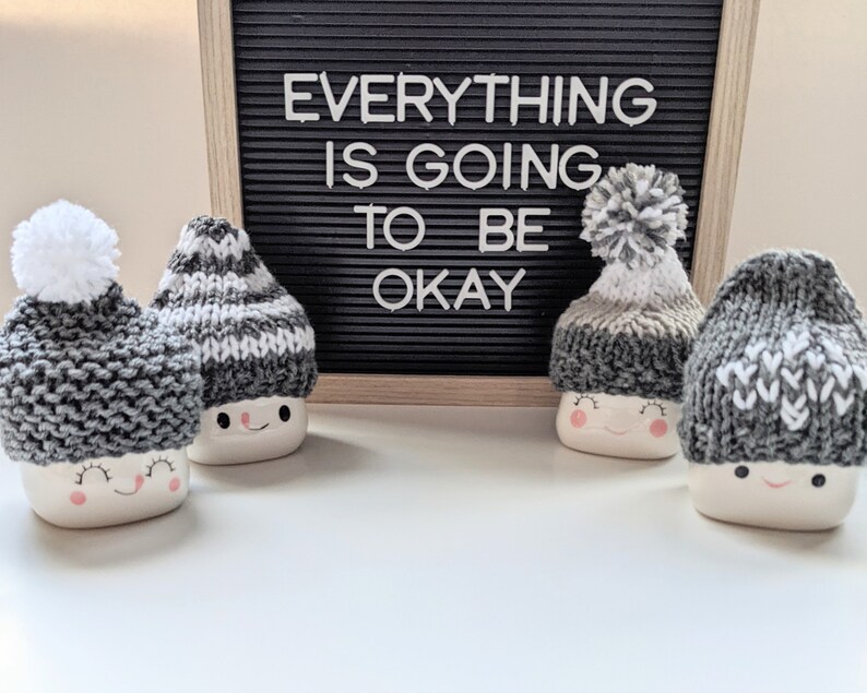 K&ouml;nnte beinhalten: Vier wei&szlig;e Keramikbecher mit fr&ouml;hlichen Gesichtern tragen gestrickte M&uuml;tzen. Die M&uuml;tzen sind grau und wei&szlig; mit Bommeln. Die Becher stehen auf einer wei&szlig;en Oberfl&auml;che vor einem schwarzen Buchstabenbrett mit der Aufschrift "Alles wird gut."