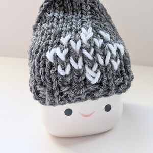 K&ouml;nnte beinhalten: Eine graue Strickm&uuml;tze mit einem wei&szlig;en Zickzackmuster. Die M&uuml;tze sitzt auf einem wei&szlig;en Keramiktopf mit einem l&auml;chelnden Gesicht.