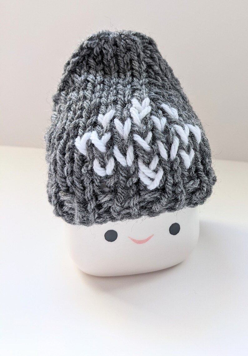 K&ouml;nnte beinhalten: Eine graue Strickm&uuml;tze mit einem wei&szlig;en Zickzackmuster. Die M&uuml;tze sitzt auf einem wei&szlig;en Keramiktopf mit einem l&auml;chelnden Gesicht.