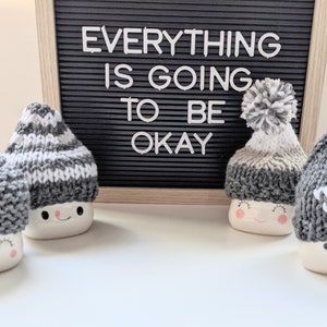K&ouml;nnte beinhalten: Vier wei&szlig;e Keramikbecher mit fr&ouml;hlichen Gesichtern, jeder mit einer anderen grauen und wei&szlig;en Strickm&uuml;tze mit Bommel. Die Becher stehen auf einer wei&szlig;en Oberfl&auml;che vor einer schwarzen Buchstabenwand mit der Aufschrift "Alles wird gut."