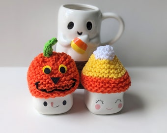 Puede incluir: Imagen con temática de Halloween con dos tazas de cerámica con caras sonrientes, cubiertas con diseños de calabaza y maíz de caramelo de ganchillo. Una taza con forma de fantasma sostiene maíz de caramelo en el fondo. La calabaza tiene un tallo verde y rasgos negros.