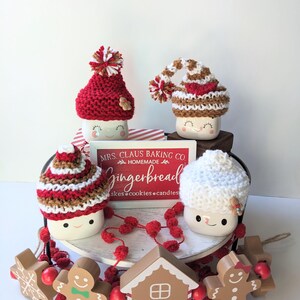 Puede incluir: Una bandeja de tres niveles decorada con un tema de pan de jengibre para Navidad. La bandeja presenta tres tazas con gorros tejidos, una casa de pan de jengibre, hombres de pan de jengibre y un letrero que dice "Mrs. Claus Baking Co. Homemade Gingerbread Cakes, Cookies, Candles".
