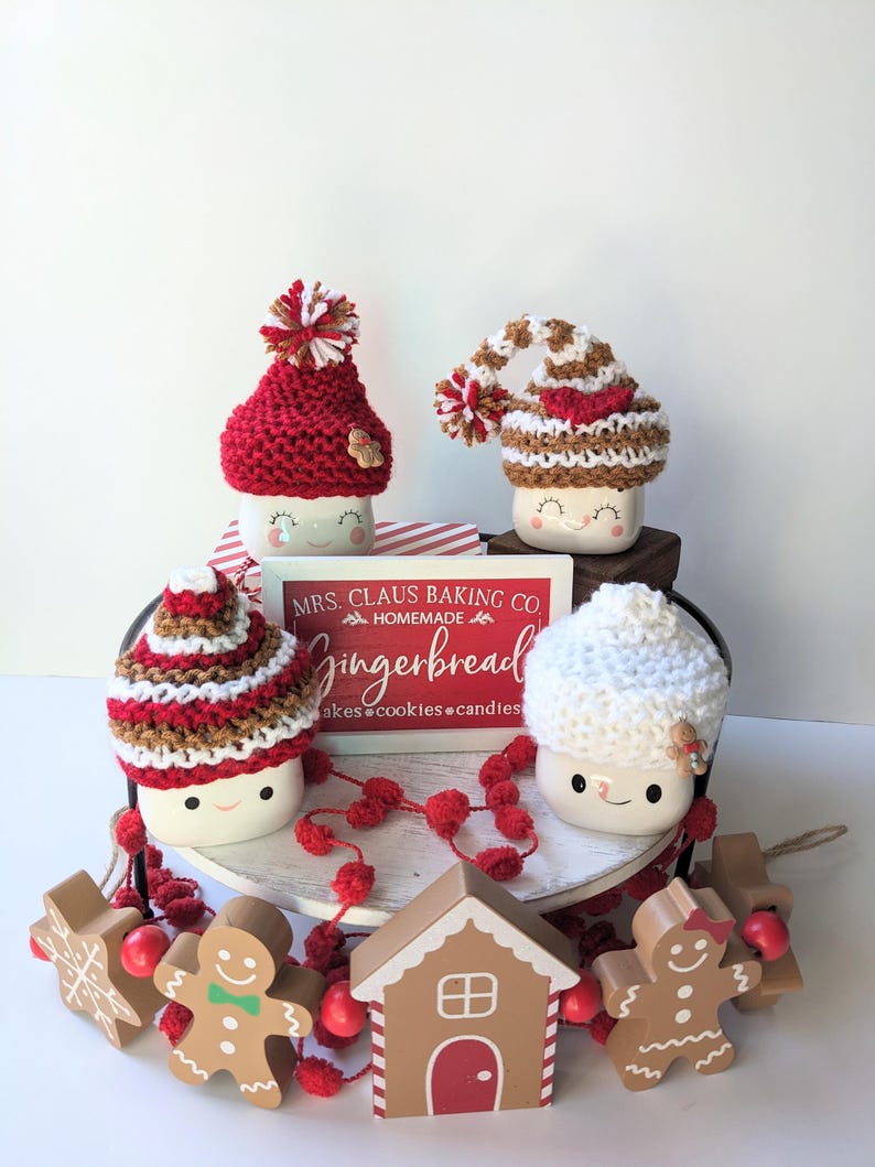 Puede incluir: Una bandeja de tres niveles decorada con un tema de pan de jengibre para Navidad. La bandeja presenta tres tazas con gorros tejidos, una casa de pan de jengibre, hombres de pan de jengibre y un letrero que dice "Mrs. Claus Baking Co. Homemade Gingerbread Cakes, Cookies, Candles".