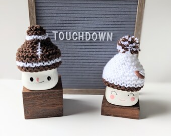 May include: Two white ceramic mugs with cute faces, wearing knitted hats. One hat is brown and white with a football design, the other is white with a brown pom-pom. The mugs are sitting on wooden blocks. A letter board behind the mugs reads "TOUCHDOWN".