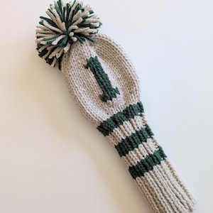 May include: A beige and dark green knitted golf club head cover featuring a large pom-pom on top and the number 1 embroidered in dark green on the front. The design includes horizontal dark green stripes on the lower section of the cover.