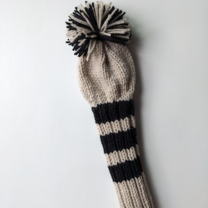 May include: A beige knitted golf club head cover with black stripes and a black and white pom pom on top.