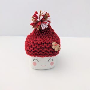 Puede incluir: Una taza de cer&aacute;mica blanca con una cara sonriente, que lleva un gorro rojo de punto con un pomp&oacute;n blanco y marr&oacute;n y un bot&oacute;n de hombre de jengibre.