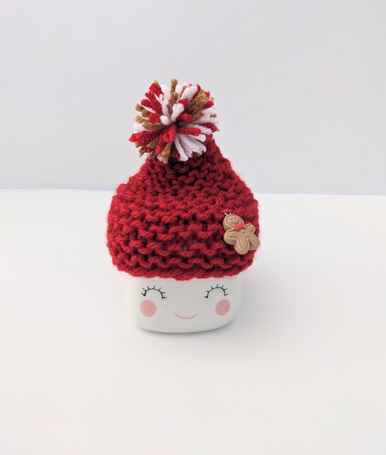 Puede incluir: Una taza de cer&aacute;mica blanca con una cara sonriente, que lleva un gorro rojo de punto con un pomp&oacute;n blanco y marr&oacute;n y un bot&oacute;n de hombre de jengibre.