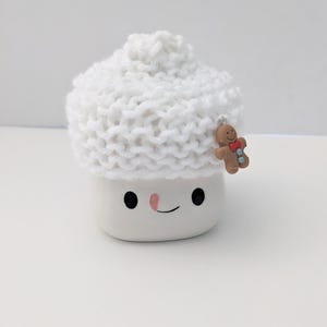 Puede incluir: Taza de cer&aacute;mica blanca con forma de malvavisco con un gorro blanco de punto y un amuleto de hombre de jengibre. La taza tiene una nariz rosa y ojos negros.