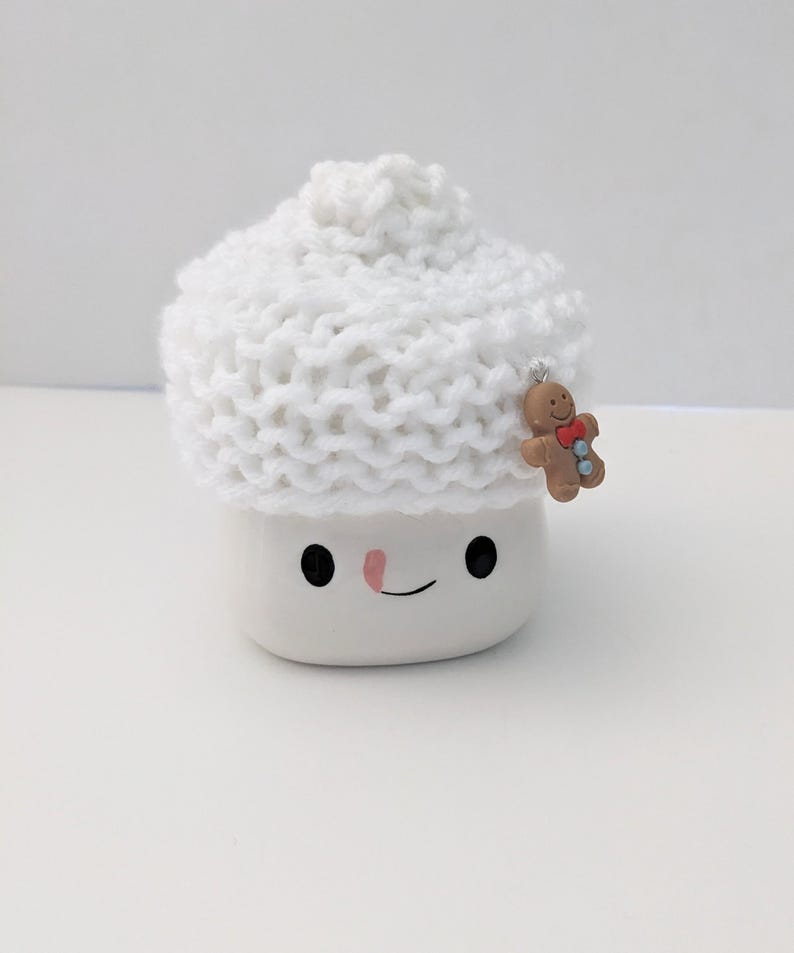 Puede incluir: Taza de cer&aacute;mica blanca con forma de malvavisco con un gorro blanco de punto y un amuleto de hombre de jengibre. La taza tiene una nariz rosa y ojos negros.