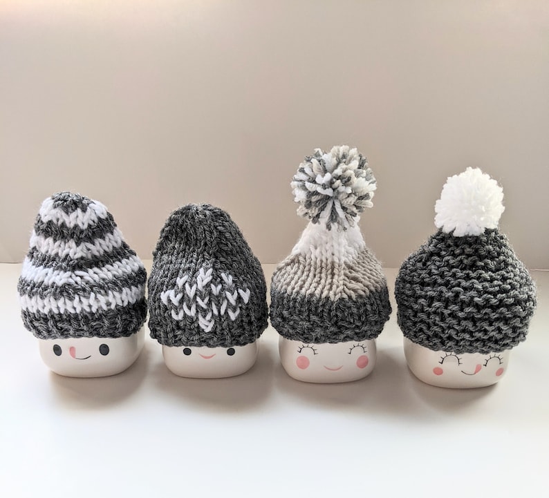 K&ouml;nnte beinhalten: Vier wei&szlig;e Keramikbecher mit grauen und wei&szlig;en Strickm&uuml;tzen. Jeder Becher hat ein anderes M&uuml;tzen-Design und ein gemaltes Smiley-Gesicht.