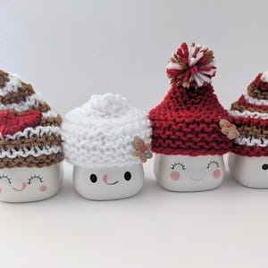 Puede incluir: Cuatro tazas de cer&aacute;mica blanca con caras sonrientes, cada una con un gorro de punto diferente. Los gorros son de rayas marrones y blancas, blancos, rojos con un pomp&oacute;n blanco y de rayas marrones y blancas con un coraz&oacute;n rojo.