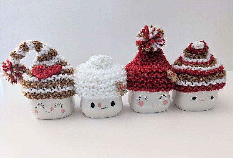 Puede incluir: Cuatro tazas de cer&aacute;mica blanca con caras sonrientes, cada una con un gorro de punto diferente. Los gorros son de rayas marrones y blancas, blancos, rojos con un pomp&oacute;n blanco y de rayas marrones y blancas con un coraz&oacute;n rojo.