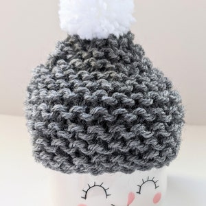 K&ouml;nnte beinhalten: Eine wei&szlig;e Keramiktasse mit einer grauen Strickm&uuml;tze und einem wei&szlig;en Bommel oben. Die Tasse hat ein l&auml;chelndes Gesicht mit geschlossenen Augen und rosa Wangen.