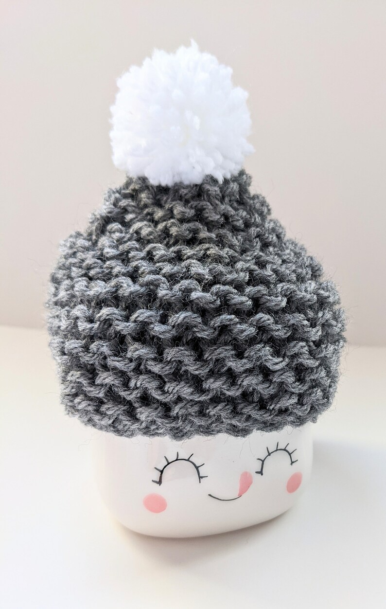 K&ouml;nnte beinhalten: Eine wei&szlig;e Keramiktasse mit einer grauen Strickm&uuml;tze und einem wei&szlig;en Bommel oben. Die Tasse hat ein l&auml;chelndes Gesicht mit geschlossenen Augen und rosa Wangen.