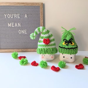 Puede incluir: Dos tazas de cerámica blanca con gorros de punto verde. Un gorro tiene una franja blanca y un corazón rojo, el otro tiene una cara verde con ojos amarillos. Las tazas están sobre una superficie blanca con pompones rojos y verdes. Un tablero de letras detrás de las tazas dice "You're a mean one."