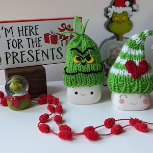 Puede incluir: Dos tazas de cerámica blanca con gorros de Grinch tejidos de color verde. Un gorro tiene un corazón rojo y el otro tiene una cara de gruñón. Las tazas están sobre una superficie blanca con una guirnalda de pompones rojos. El letrero dice "Estoy aquí por los regalos".