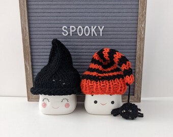 May include: Two white ceramic mugs with smiling faces, each wearing a knitted hat. One hat is black, the other is orange and black striped. A small black spider hangs from the orange hat. The word "SPOOKY" is displayed on a gray letter board.