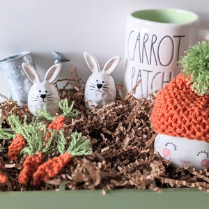 May include: An Easter-themed display featuring ceramic bunny figurines, crocheted carrots, and a mug with "CARROT PATCH" text. A ceramic figure wears an orange carrot hat with a green pom-pom. The items are arranged in a green tray with paper filler.