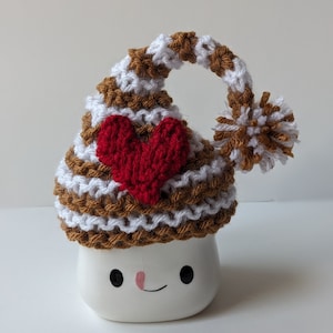 Puede incluir: Una taza de cer&aacute;mica blanca con forma de malvavisco con un gorro de punto a rayas marrones y blancas con un coraz&oacute;n rojo en la parte superior. La taza tiene una cara sonriente.