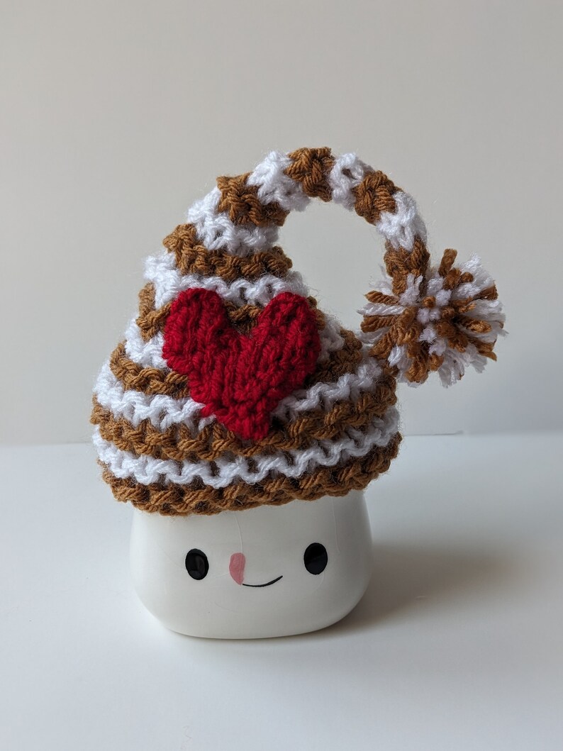 Puede incluir: Una taza de cer&aacute;mica blanca con forma de malvavisco con un gorro de punto a rayas marrones y blancas con un coraz&oacute;n rojo en la parte superior. La taza tiene una cara sonriente.