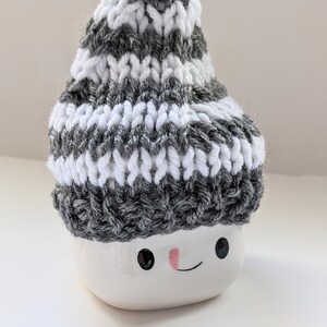 K&ouml;nnte beinhalten: Eine wei&szlig;e Keramik-Schneemann-Tasse mit einer grau-wei&szlig; gestreiften Strickm&uuml;tze. Die Tasse hat schwarze Augen und eine rosafarbene Nase.