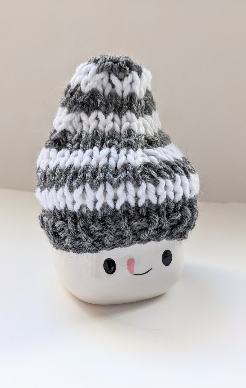 K&ouml;nnte beinhalten: Eine wei&szlig;e Keramik-Schneemann-Tasse mit einer grau-wei&szlig; gestreiften Strickm&uuml;tze. Die Tasse hat schwarze Augen und eine rosafarbene Nase.