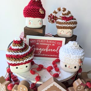 Puede incluir: Tres tazas de cer&aacute;mica blanca con caras sonrientes, cada una con un gorro de punto diferente. Los gorros son de rayas rojas y blancas, de rayas marrones y blancas y blancos. Las tazas est&aacute;n sobre un soporte de madera de varios niveles con pompones rojos y un cartel que dice "MRS. CLAUS BAKING CO. HOMEMADE Gingerbread cakes cookies candies - "