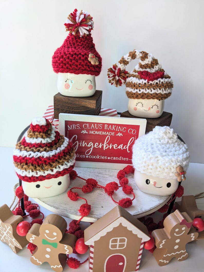 Puede incluir: Tres tazas de cer&aacute;mica blanca con caras sonrientes, cada una con un gorro de punto diferente. Los gorros son de rayas rojas y blancas, de rayas marrones y blancas y blancos. Las tazas est&aacute;n sobre un soporte de madera de varios niveles con pompones rojos y un cartel que dice "MRS. CLAUS BAKING CO. HOMEMADE Gingerbread cakes cookies candies - "