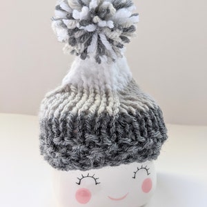 K&ouml;nnte beinhalten: Eine wei&szlig;e Keramiktasse mit einer grau-wei&szlig;en Strickm&uuml;tze mit Bommel. Die Tasse hat ein l&auml;chelndes Gesicht mit rosa Wangen und schwarzen Wimpern.