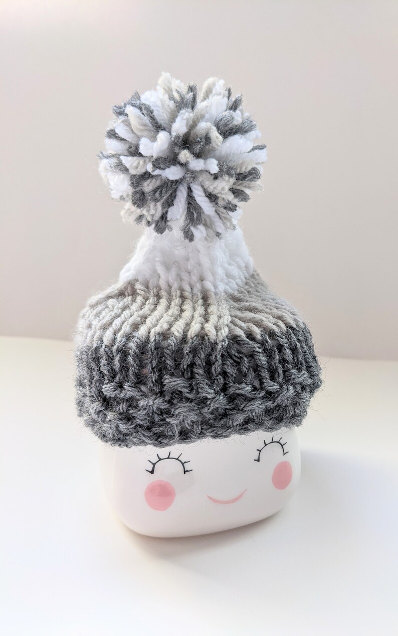 K&ouml;nnte beinhalten: Eine wei&szlig;e Keramiktasse mit einer grau-wei&szlig;en Strickm&uuml;tze mit Bommel. Die Tasse hat ein l&auml;chelndes Gesicht mit rosa Wangen und schwarzen Wimpern.
