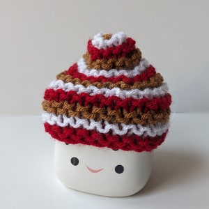 Puede incluir: Una taza de cer&aacute;mica blanca con una cara sonriente, que lleva un gorro de punto con rayas rojas, blancas y marrones.